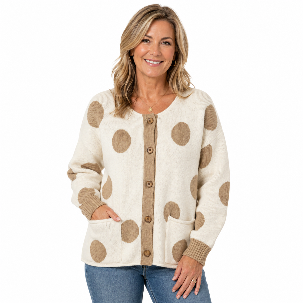 A smiling woman wearing a cream knit cardigan with large tan polka dots and a tan button placket, paired with blue jeans, against a plain white background.