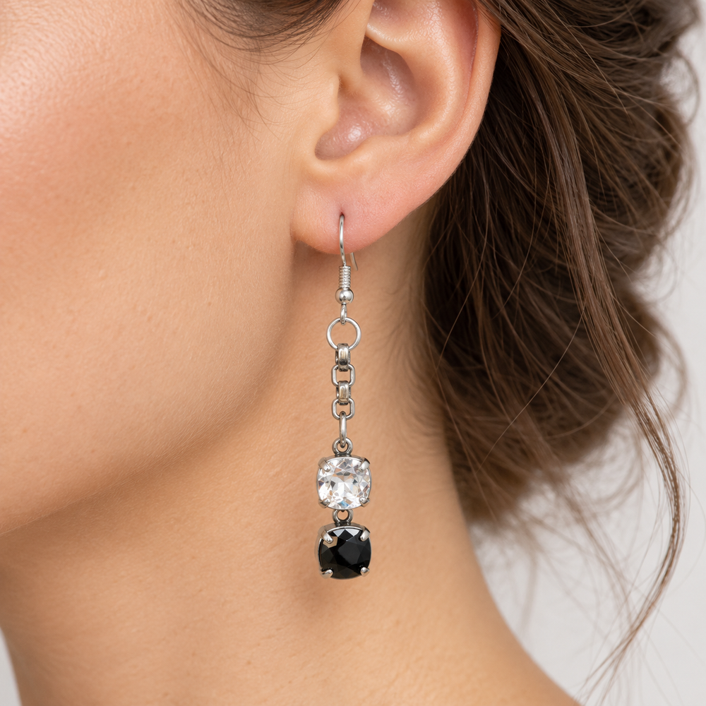 A close-up of a model's ear wearing a silver drop earring with a clear round gemstone and a black square gemstone.