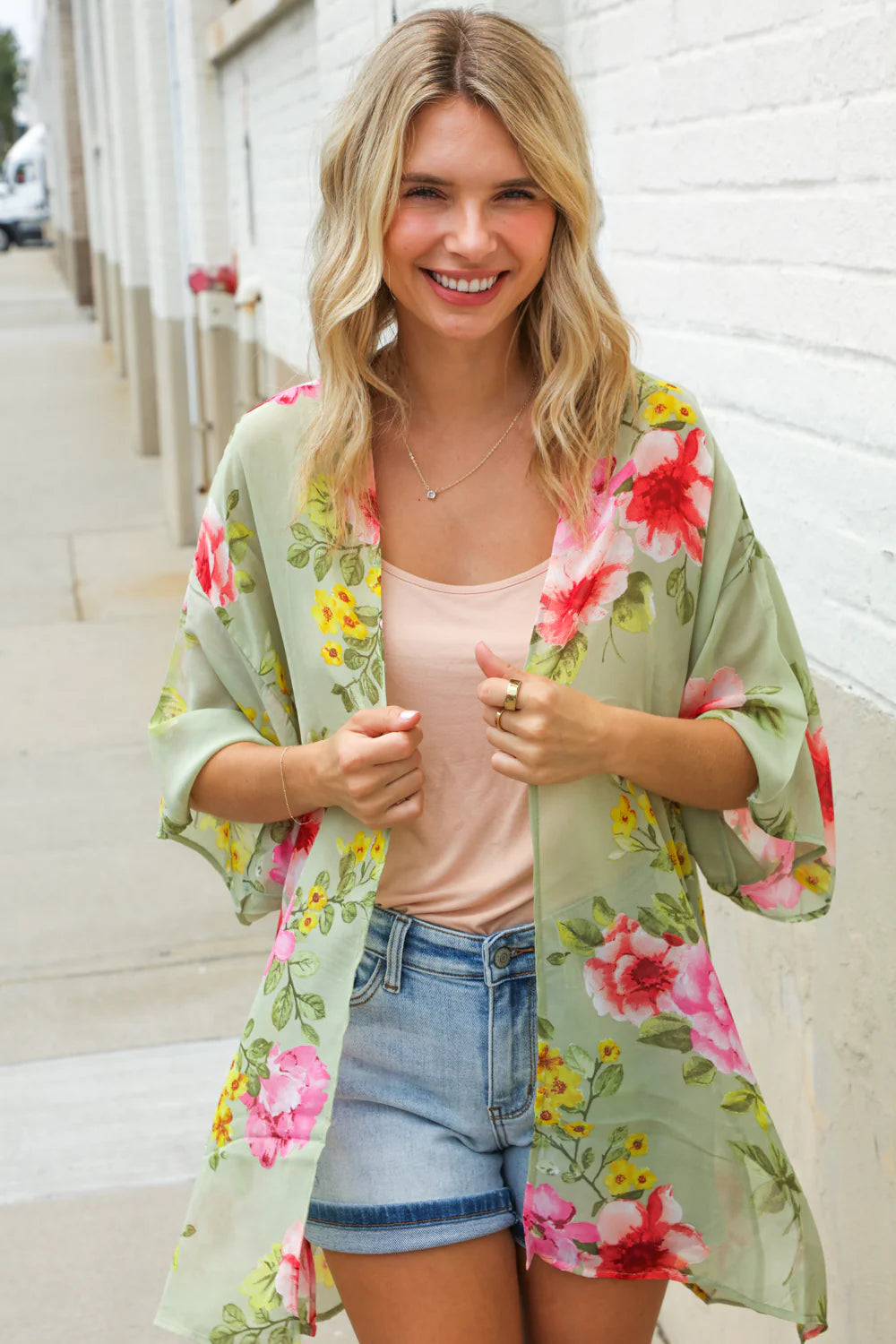 Woman wearing a floral kimono over a pink top and denim shorts, standing against a white wall.