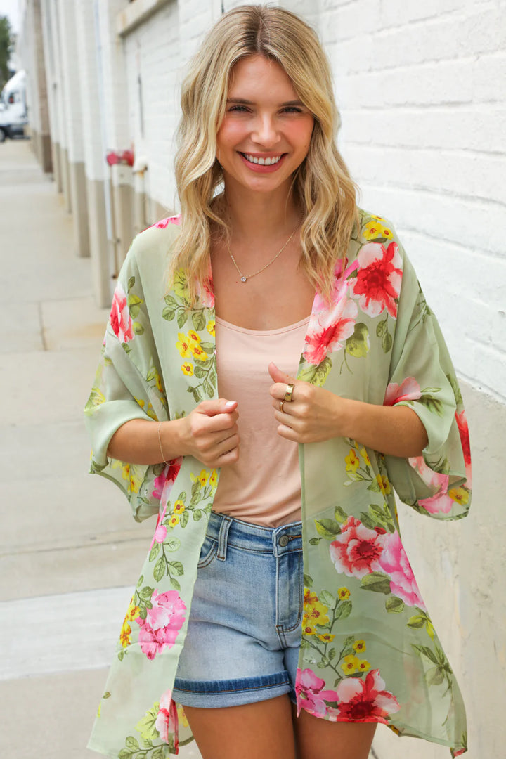 Woman wearing a floral kimono over a pink top and denim shorts, standing against a white wall.
