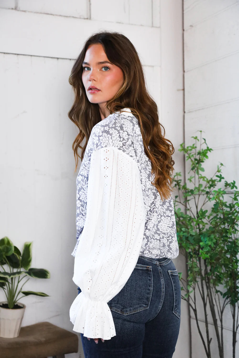 Woman wearing a white lace top and blue jeans standing indoors with plants in the background