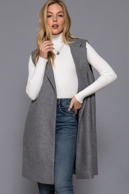 Woman wearing a long gray vest over a white sweater and blue jeans on a gray background