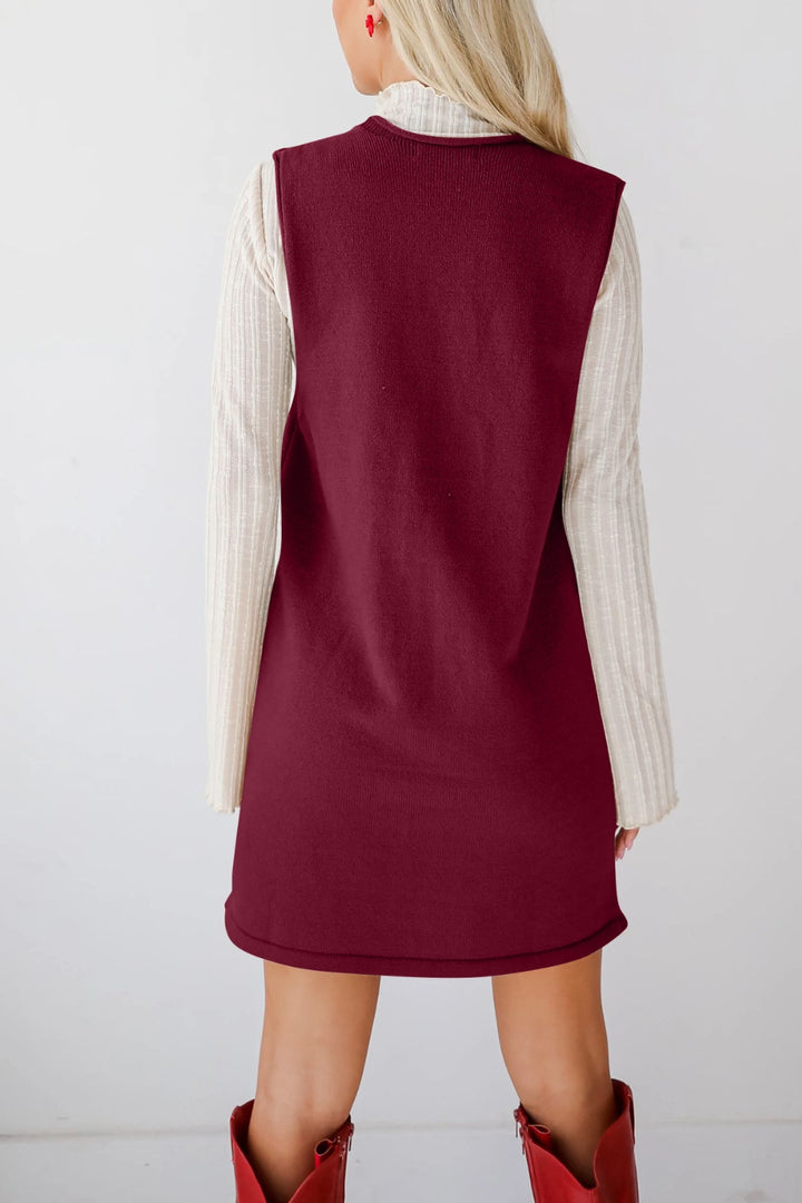 Maroon sleeveless dress worn over a white sweater with red boots on a plain background
