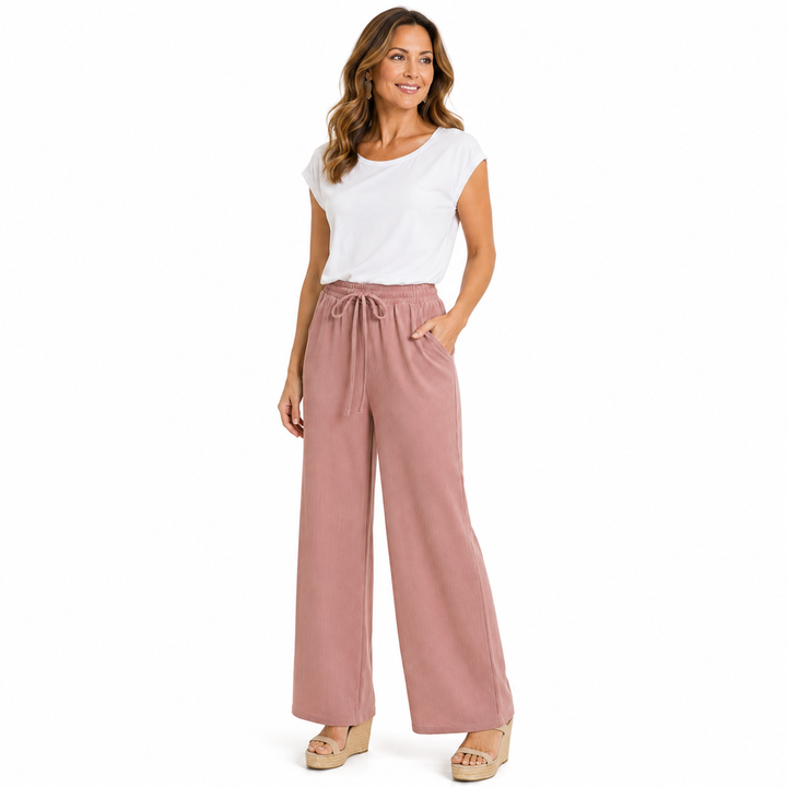 A woman modeling a pair of dusty rose wide-leg pants with a drawstring waist, paired with a white t-shirt and beige wedge sandals, against a plain white background.