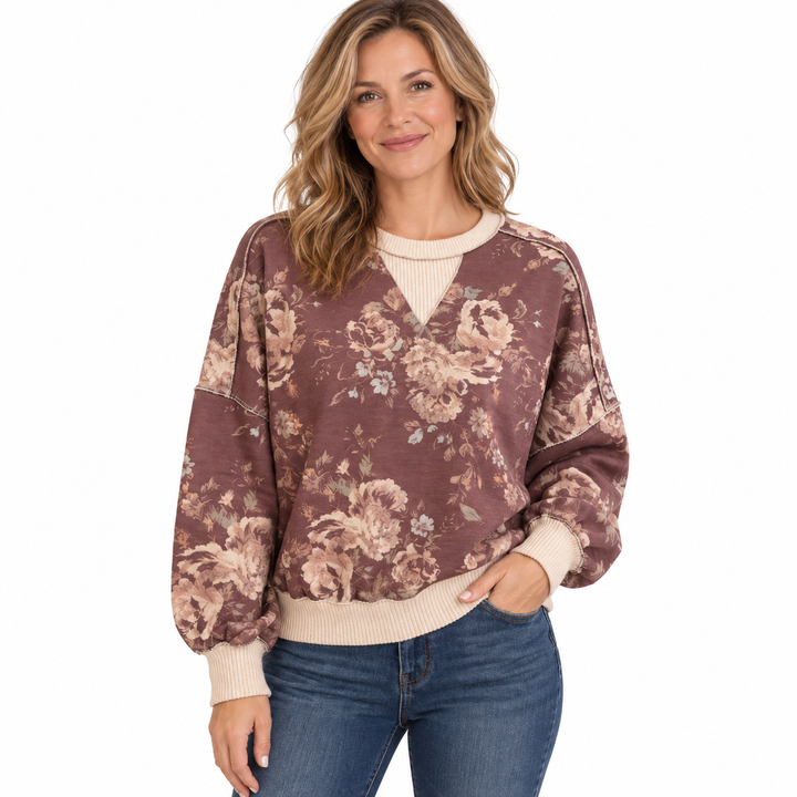 A woman modeling a brown and beige floral patterned sweatshirt with ribbed cream trim, paired with blue jeans, against a plain white background.