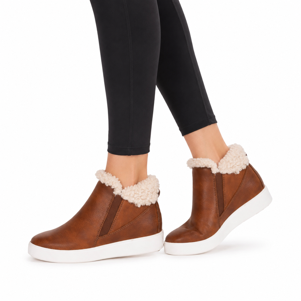 A person wearing brown ankle slipper boots with cream sherpa lining and white platform soles, paired with black leggings, shown against a plain white background.