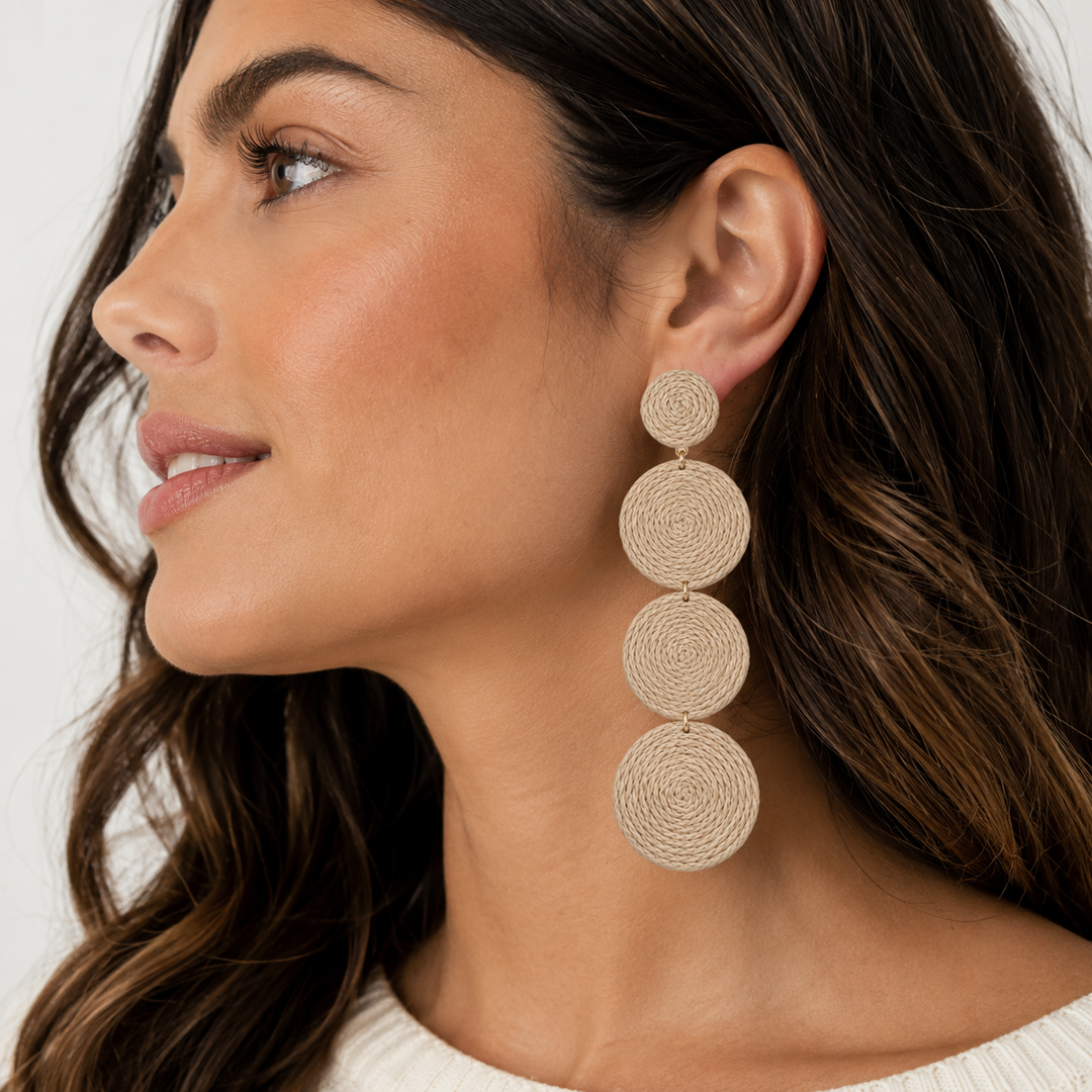 A close-up of a woman wearing a long, dangling statement earring made of three stacked, circular, woven beige discs.