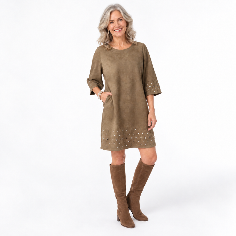 A smiling woman stands against a clean white studio background wearing a fatigue green suede-like dress with elbow-length sleeves, side pockets, and small metallic stud embellishments near the cuffs and hem, paired with matching brown knee-high boots.