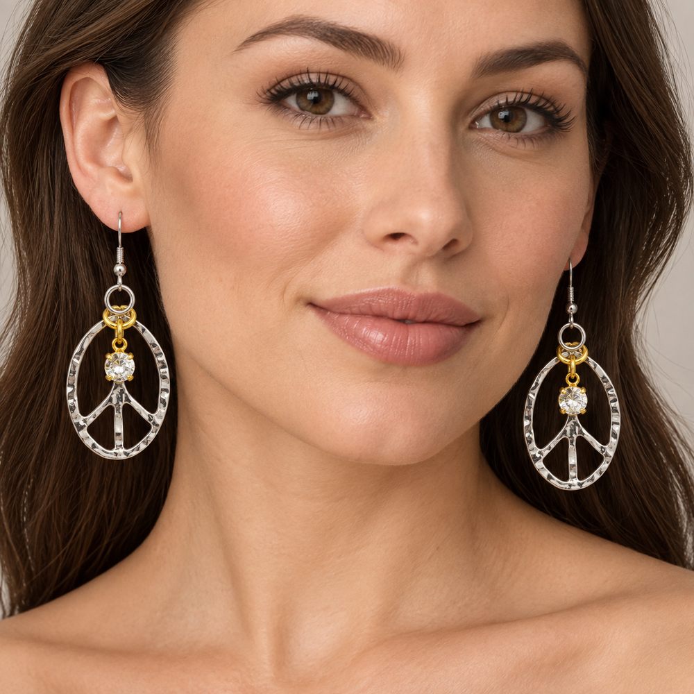 A close-up of a woman wearing silver peace sign dangle earrings with gold accents and clear rhinestones, set against a soft neutral background.