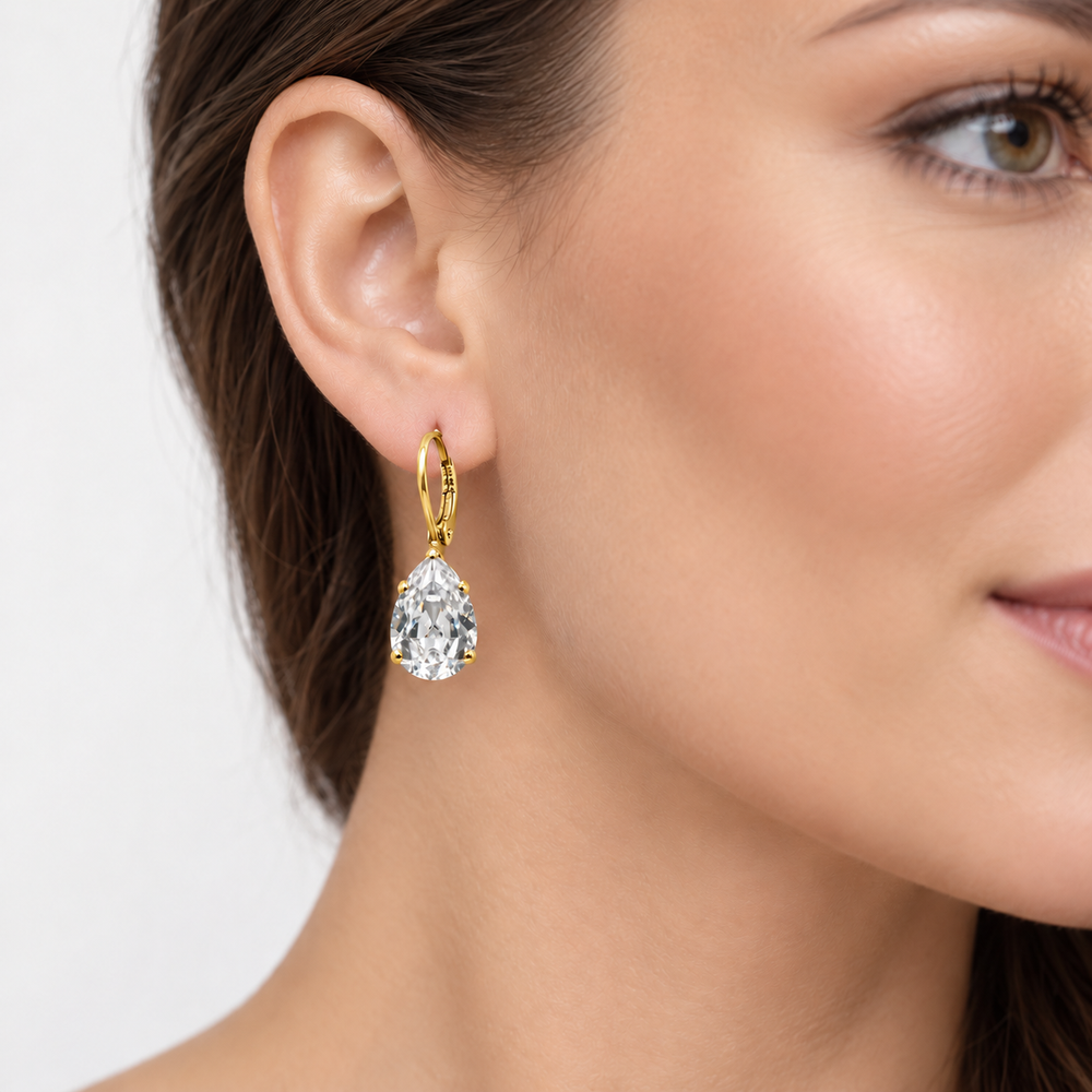 A close-up of a woman's ear wearing a gold drop earring with a teardrop-shaped clear gemstone, set against a clean light gray background.