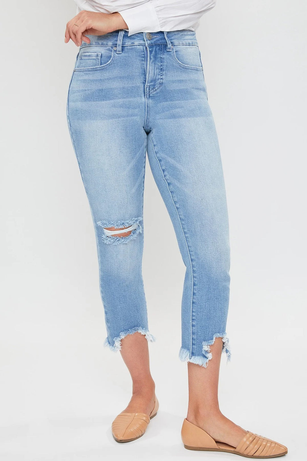 A woman sitting on a bench wearing a white shirt and blue distressed crop jeans with frayed hem, paired with beige strappy sandals.