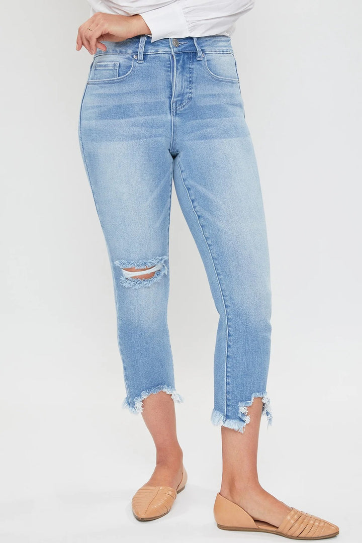 A woman sitting on a bench wearing a white shirt and blue distressed crop jeans with frayed hem, paired with beige strappy sandals.