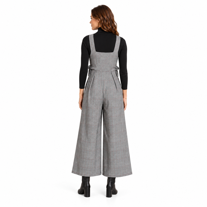 A woman viewed from the back wearing a gray wide-leg plaid jumpsuit over a black long-sleeve top and black ankle boots, against a plain white studio background.