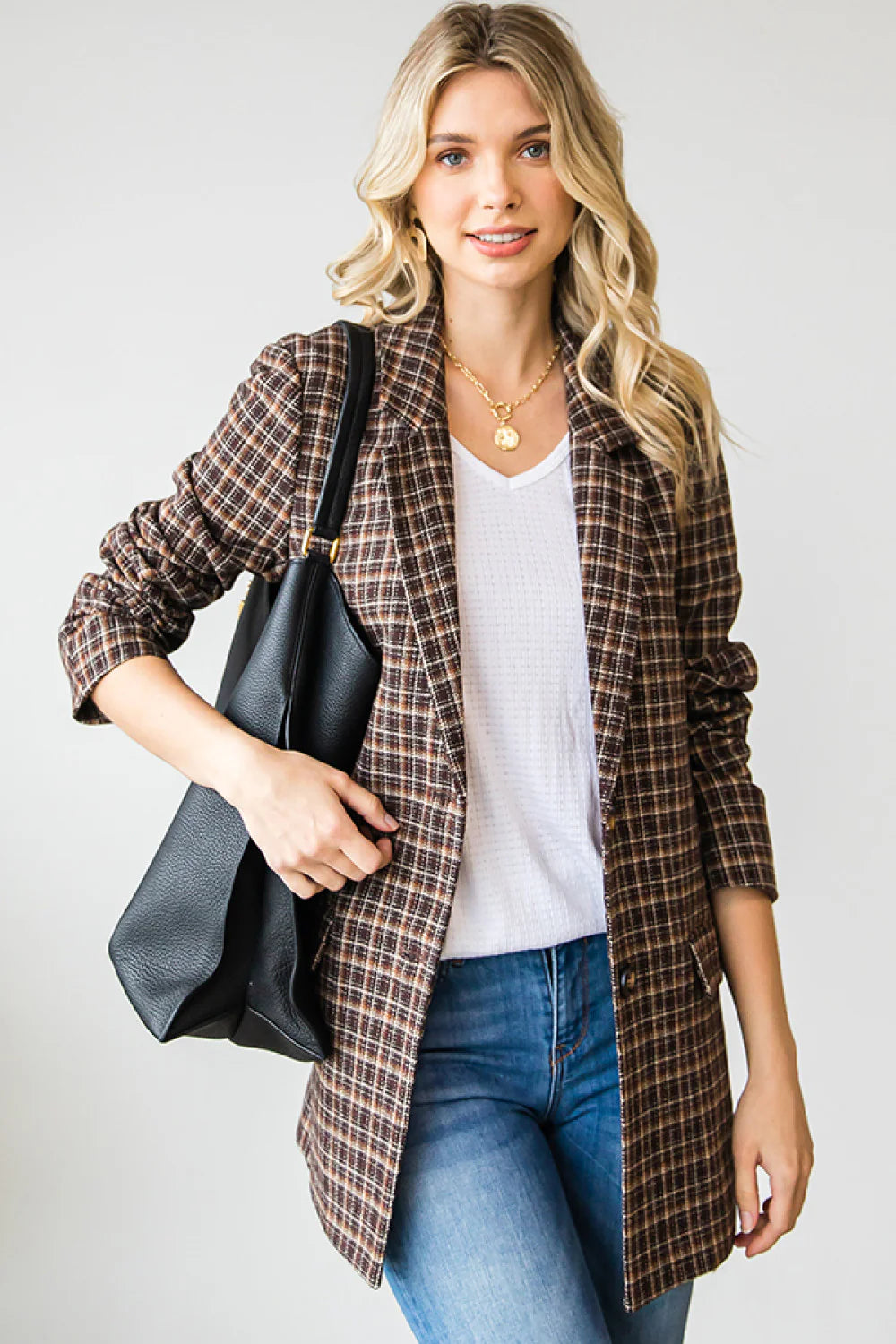 Woman wearing a plaid jacket, white shirt, and blue jeans, holding a black bag on a plain background