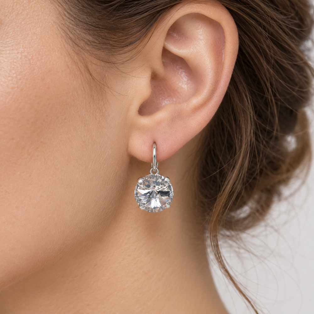 A close-up of a woman's ear wearing a single dangling stud earring with a clear faceted gemstone in a silver-toned setting.