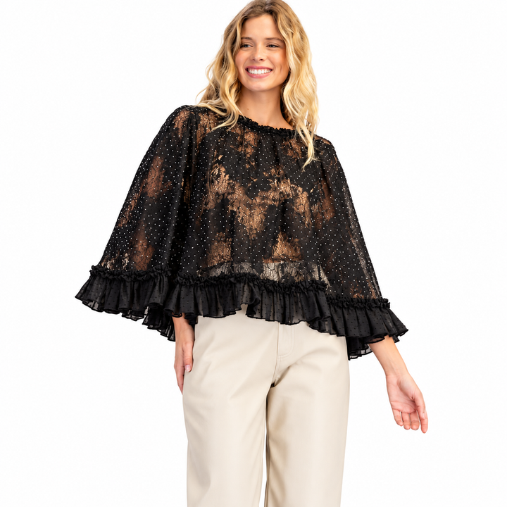 A woman modeling a sheer black lace top with wide flutter sleeves and ruffled trim, paired with light beige pants, against a plain white studio background.