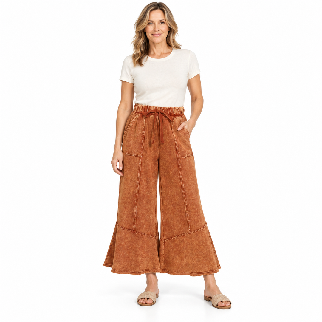 A woman modeling a pair of rust-brown wide-leg cropped pants with a drawstring waist, paired with a white t-shirt and beige sandals, against a plain white background.
