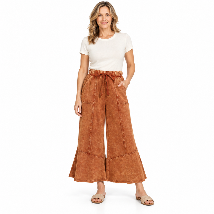 A woman modeling a pair of rust-brown wide-leg cropped pants with a drawstring waist, paired with a white t-shirt and beige sandals, against a plain white background.