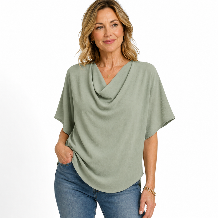 A woman modeling a sage green cowl neck short-sleeve top with blue jeans against a plain white background.