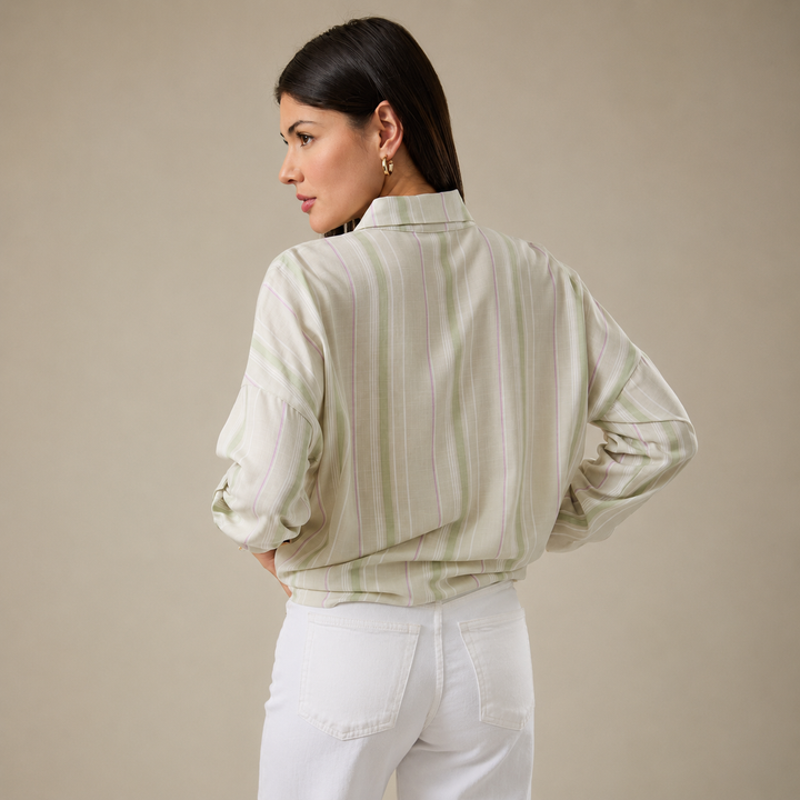A woman shown from the back wearing a light sage green and white vertically striped long-sleeve blouse with white pants, set against a solid beige studio background.