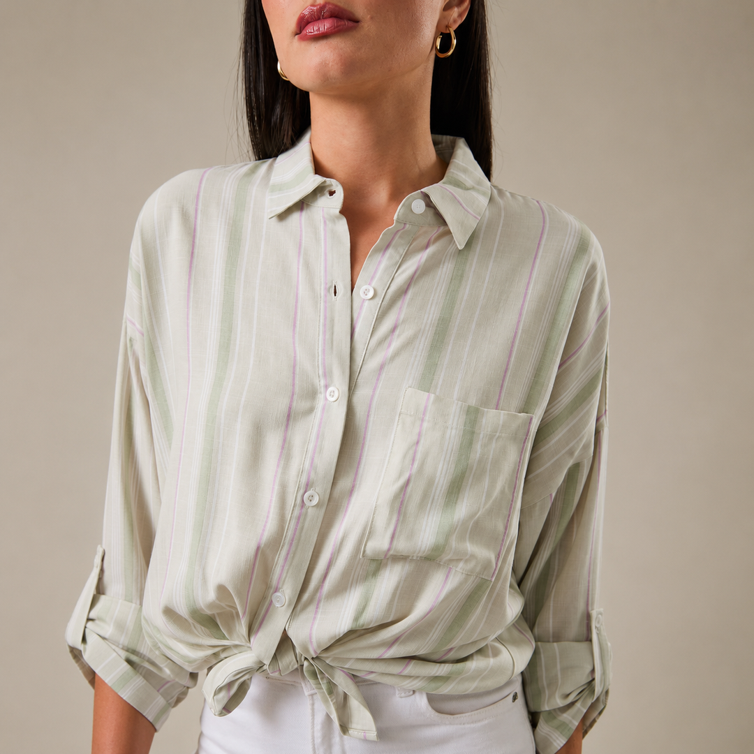 A woman wearing a light sage green and white vertically striped button-down shirt with a chest pocket, styled with a knot at the waist and white pants against a neutral beige background.
