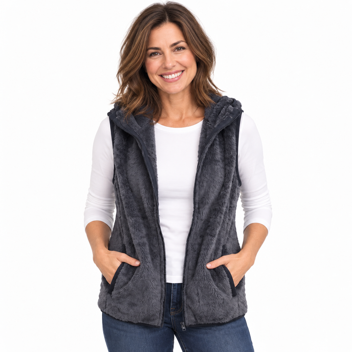 A smiling woman wearing a dark gray fuzzy hooded zip-front vest over a white long-sleeve top and blue jeans, standing against a plain white background.