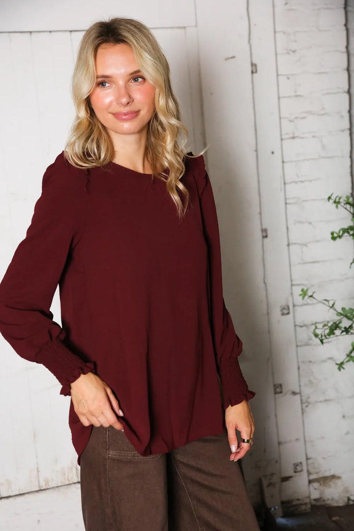 Woman wearing a burgundy top and brown pants against a white wooden wall.