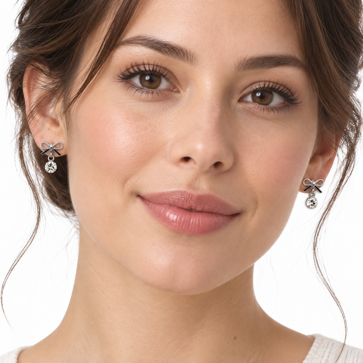 A close-up of a woman wearing small bow-shaped stud earrings with dangling clear gemstones, shown against a plain white background.