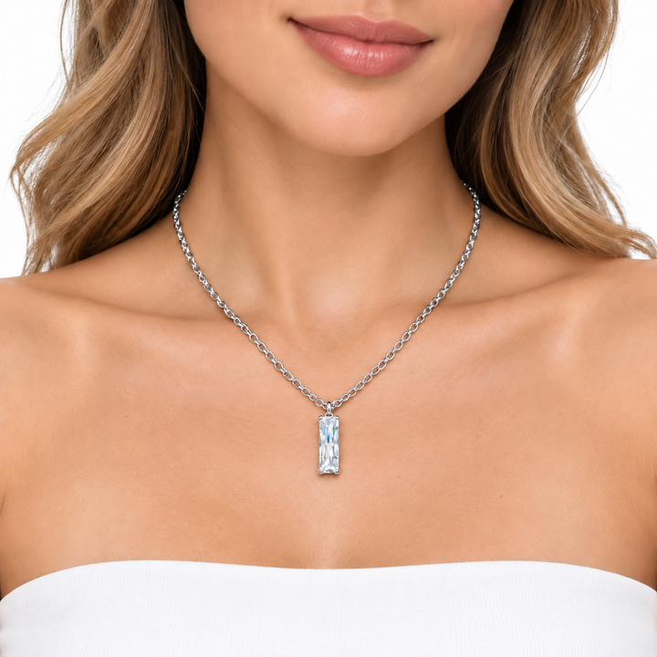 A close-up of a woman wearing a silver-toned chain necklace with a rectangular pendant featuring a textured, iridescent blue and white pattern, set against a clean white background.