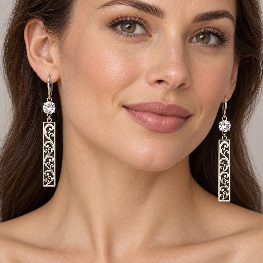 A close-up of a woman wearing a pair of silver-toned drop earrings with round clear stones and elongated filigree rectangular dangles, shown against a soft neutral background.