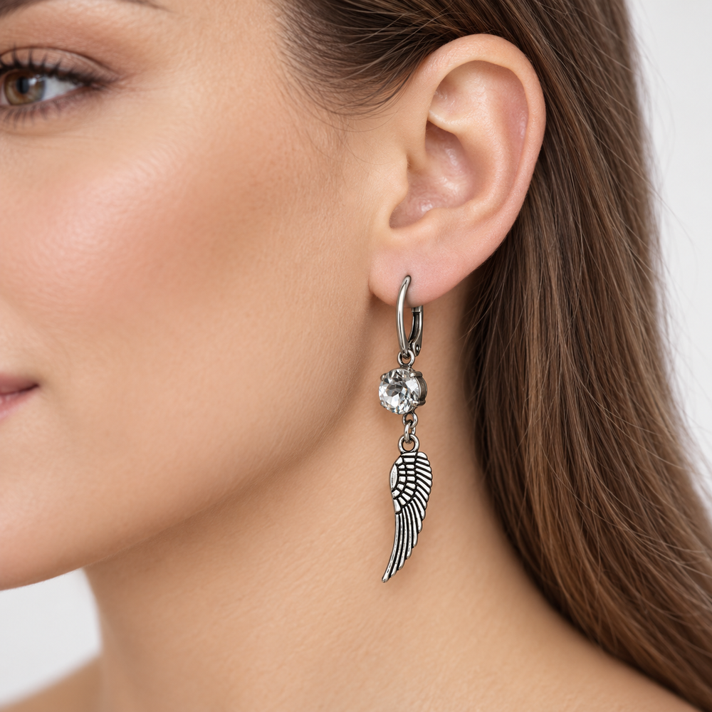 A close-up of a woman's ear wearing a silver hoop earring with a dangling wing charm and a clear faceted crystal.