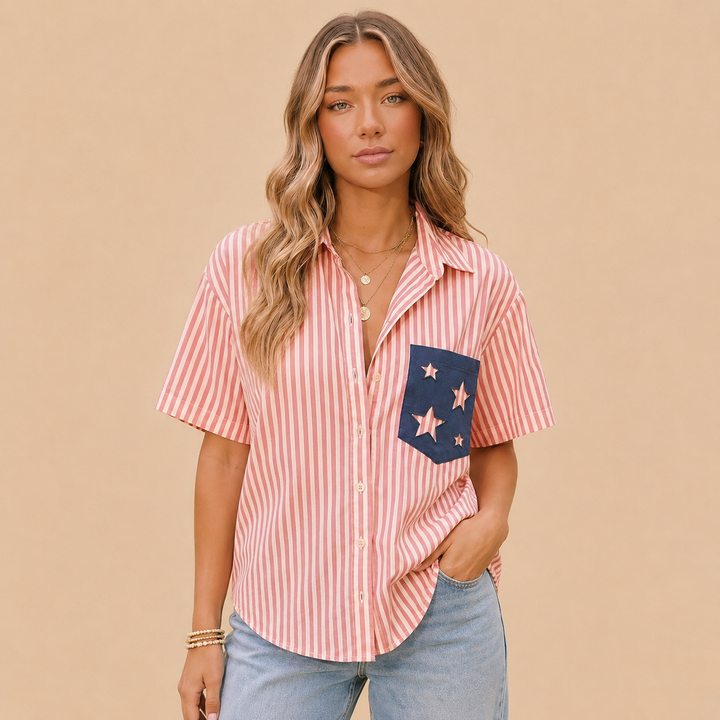 A woman modeling a short-sleeve button-up shirt with pink and white vertical stripes, featuring a navy blue pocket on the left chest decorated with pink stars, paired with light blue jeans against a solid beige background.