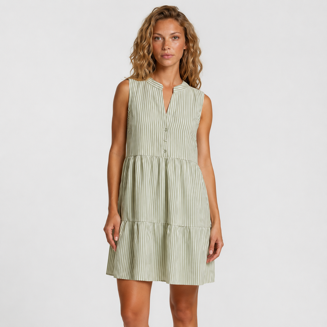 A woman modeling a sleeveless tiered mini dress with vertical stripes in sage green and white, set against a plain light gray studio background.