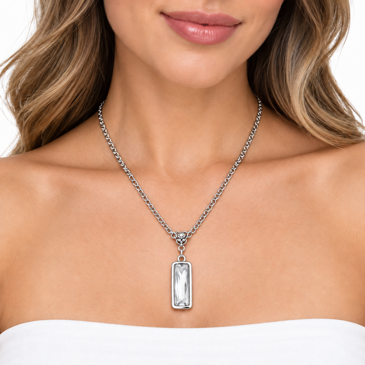 A close-up of a woman wearing a silver-toned chain necklace with a rectangular clear gemstone pendant, shown against her collarbone and chest.