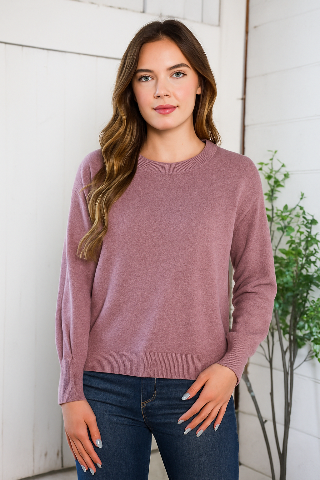Woman wearing a mauve sweater and dark jeans against a white wall.