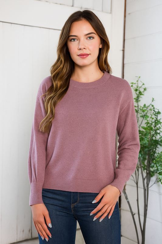 Woman wearing a mauve sweater and dark jeans against a white wall.