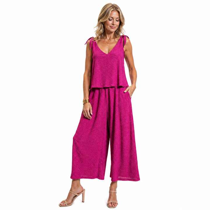 A woman modeling a bright magenta jumpsuit with tie details at the shoulders shown against a plain white background.