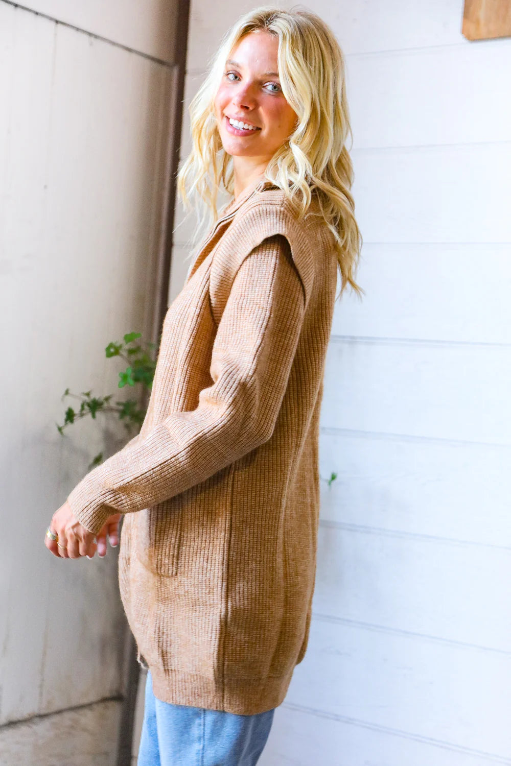 Woman wearing a brown cardigan standing against a white wall.