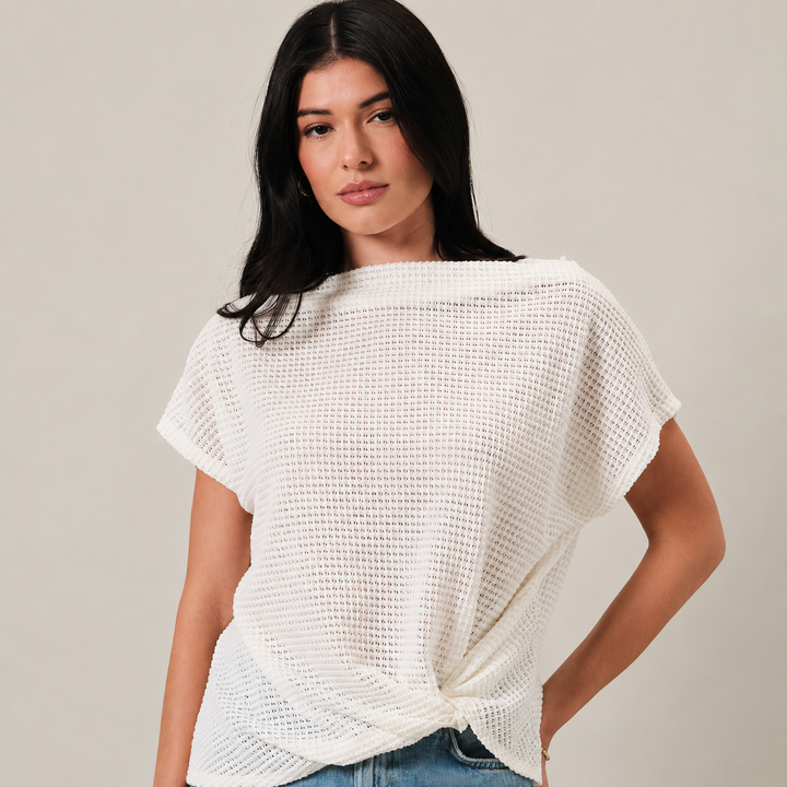 A woman wearing a white waffle-knit short-sleeve top with a twisted hem, paired with blue jeans, against a solid light beige background.