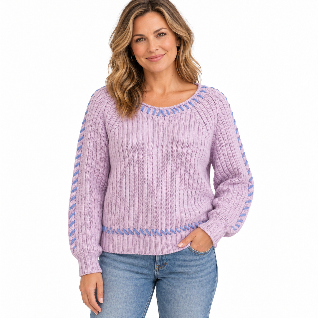 A woman wearing a lavender ribbed knit sweater with blue patterned trim along the neckline, cuffs, and hem, paired with blue jeans, against a plain white background.