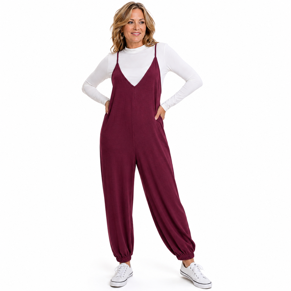 A woman modeling a burgundy sleeveless jumpsuit over a white long-sleeve mock neck top, paired with white sneakers, on a solid white background.