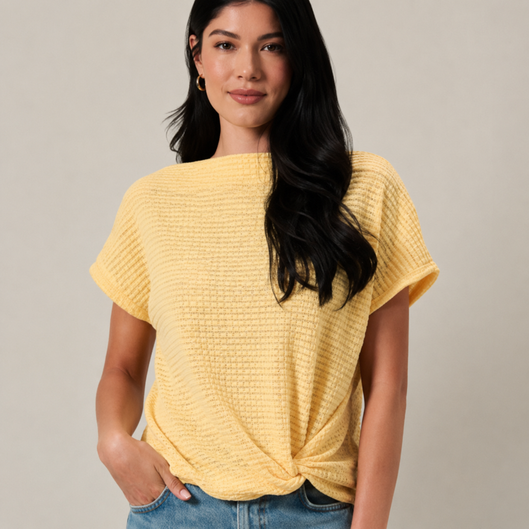 A woman wearing a pale yellow waffle-knit short-sleeve top with a twisted hem, paired with blue jeans, against a solid light gray background.