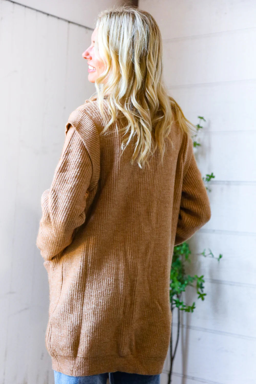 Person wearing a brown cardigan standing against a white wall with greenery.