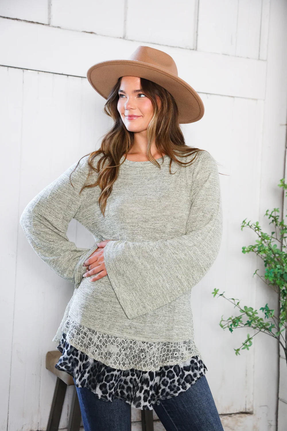 Woman wearing a long, light gray sweater with a leopard print hem, standing against a white brick wall.