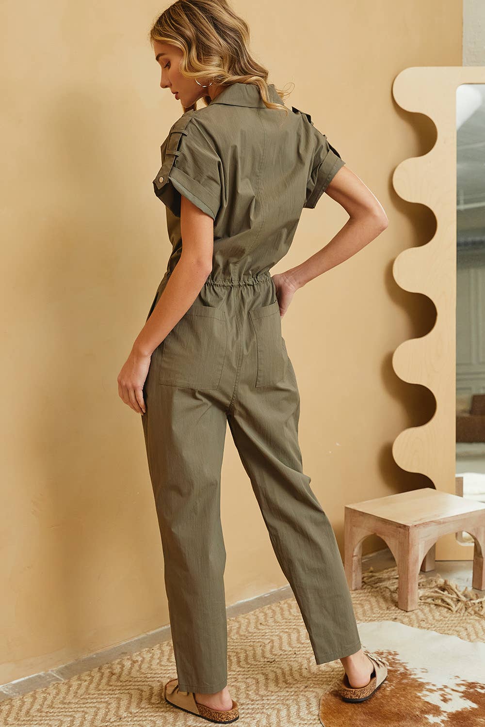 Woman wearing a green jumpsuit in a room with a beige wall and wooden floor.
