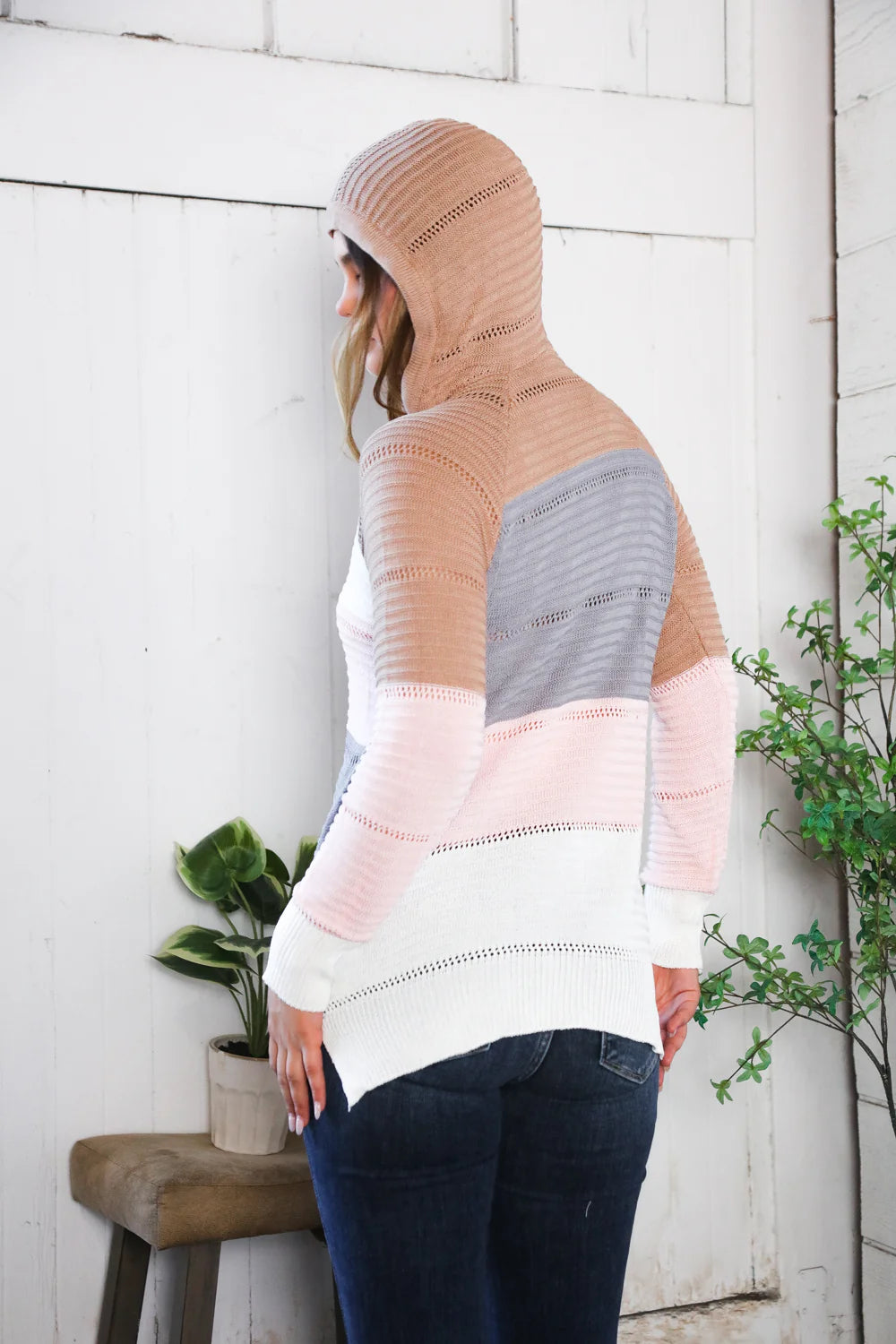 Person wearing a hooded sweater standing against a white wooden wall with plants.