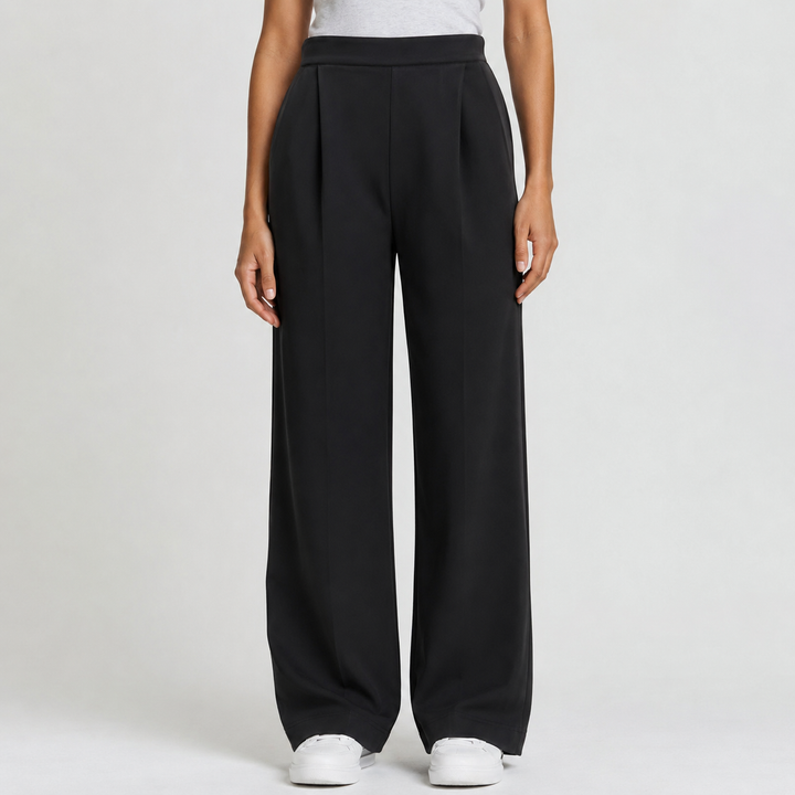 A model wearing black wide-leg pleated trousers with a matching black belt, paired with a light gray top and white sneakers, shown from the waist down on a light gray studio background.