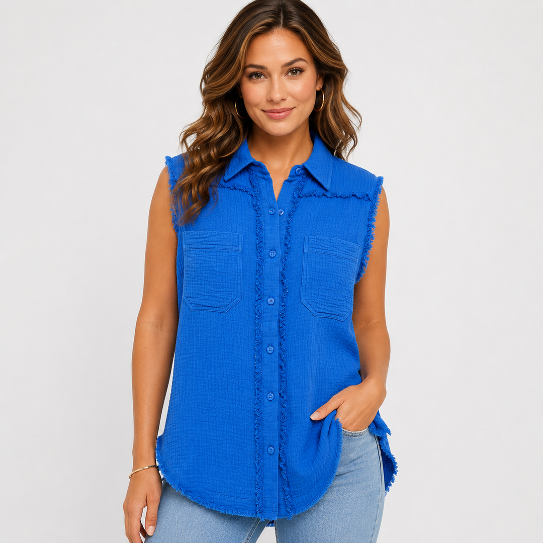 A woman modeling a sleeveless bright blue textured button-up shirt with frayed edges, two chest pockets, and a curved hem, paired with light blue jeans against a light gray studio background.