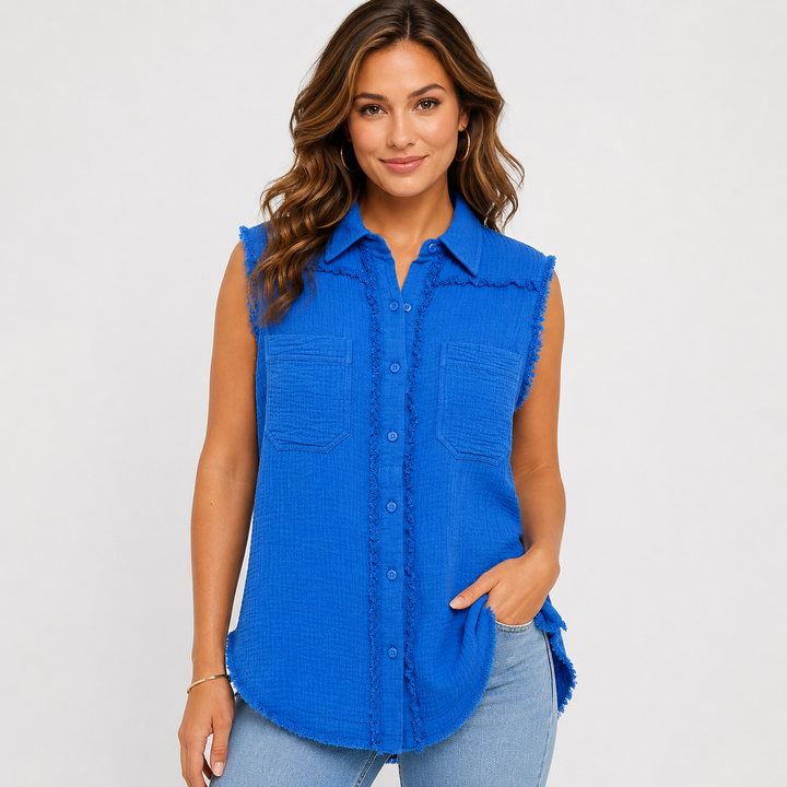 A woman modeling a sleeveless bright blue textured button-up shirt with frayed edges, two chest pockets, and a curved hem, paired with light blue jeans against a light gray studio background.