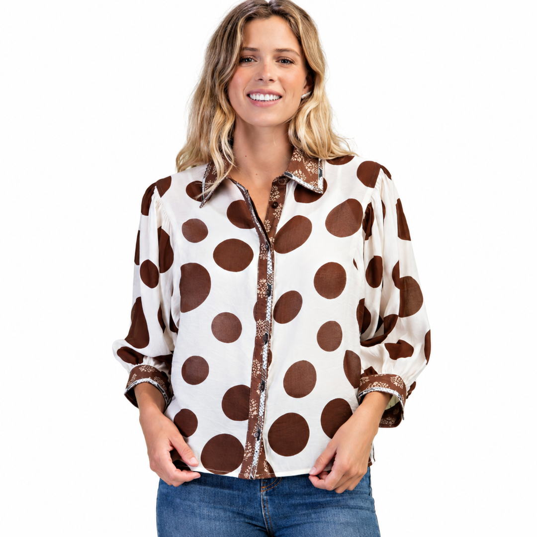 A woman wearing a long-sleeve button-up blouse with a brown and cream polka dot pattern and patterned trim, paired with blue jeans, against a plain white background.